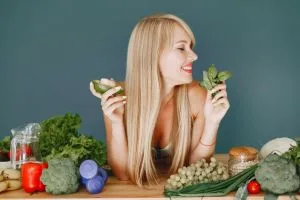 A smiling woman holding avocado and basil, surrounded by fresh vegetables and fr