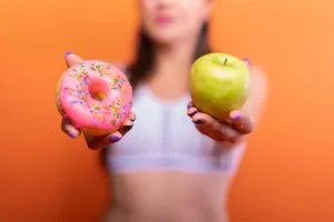 A woman holds a green apple and a pink donut with sprinkles against an orange ba