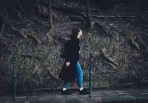 A woman in a long coat walks on a sidewalk surrounded by exposed tree roots.