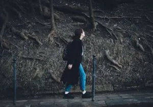 A woman in a long coat walks on a sidewalk surrounded by exposed tree roots.